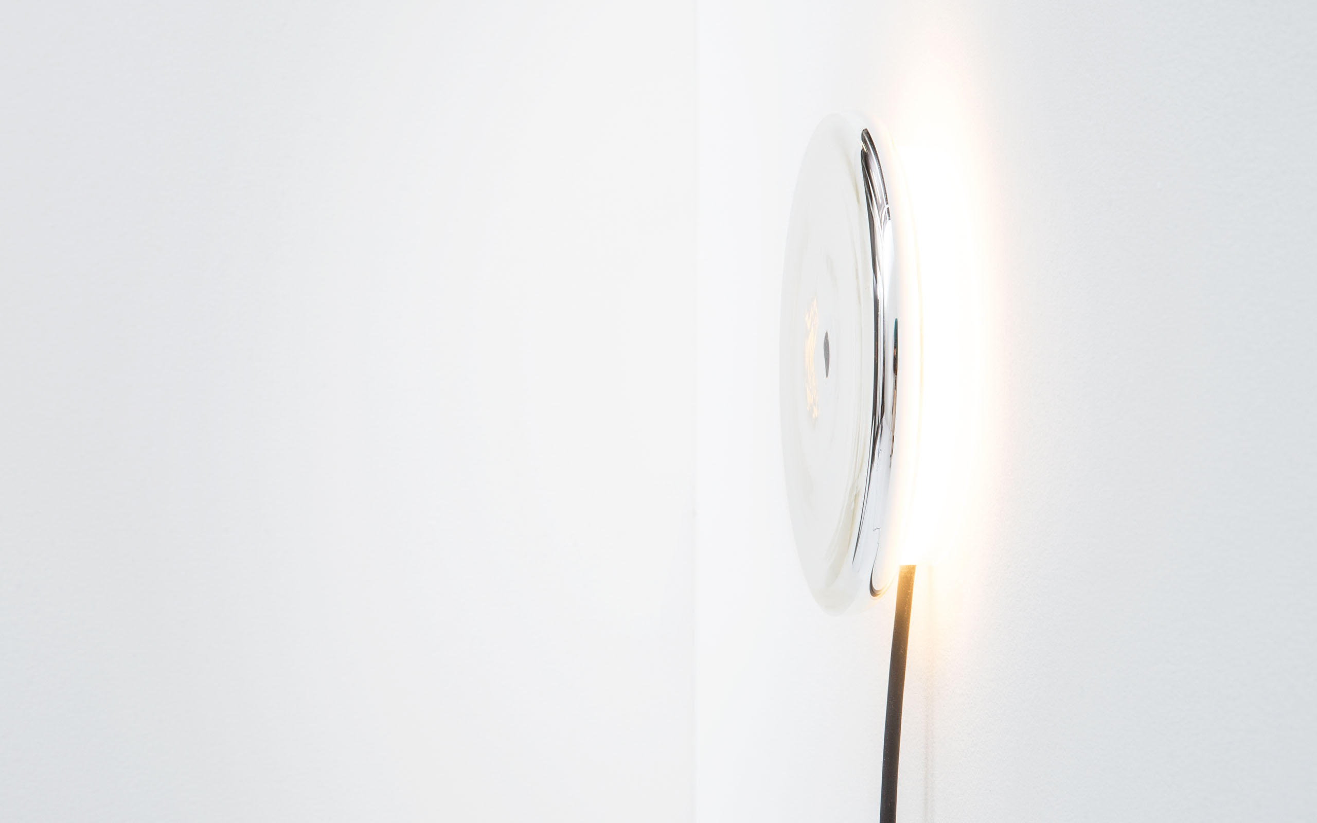 A wall light is shown from the side. The body of the light, a silver plated disc reflects the light emitted behind it.