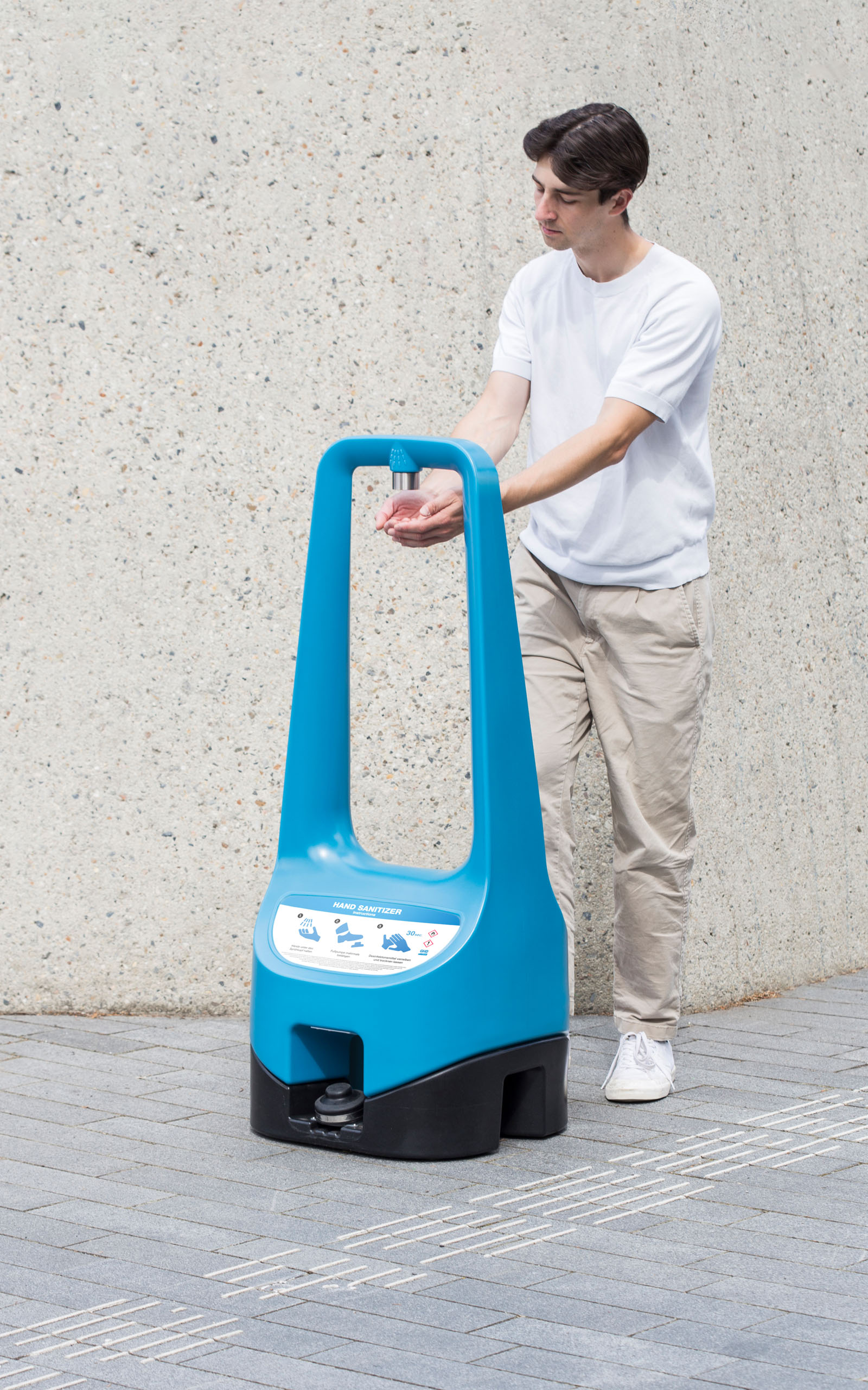 A person shown from the front is using a foot pump to enable a Toi Care hand sanitizer standing outdoors.